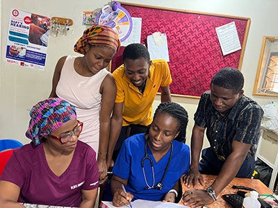 A group of medical professionals surrounds a table. A female doctor in blue scrubs writes on a document, while others attentively observe. Collaborative atmosphere.