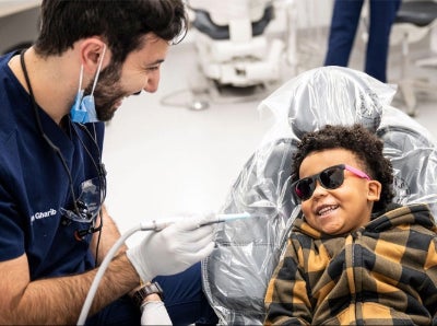 A dentist in scrubs and gloves smiles at a child wearing sunglasses and a plaid jacket, sitting in a dental chair, creating a joyful, relaxed atmosphere.