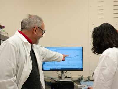 Two researchers in lab coats look at a computer monitor displaying a graph, while one points at the screen as they discuss the data.