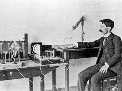 A man in a suit sits at a table operating early radio equipment with wires and bulbs, exuding focus and curiosity in a historical laboratory setting.
