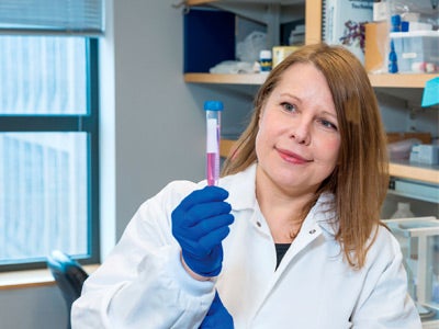 A scientist in a lab coat and blue gloves holds a test tube with pink liquid, examining it thoughtfully in a laboratory setting.