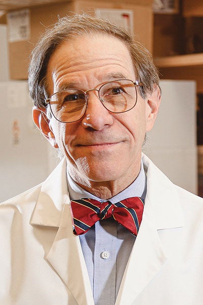 Older adult male in glasses and a white lab coat, smiling warmly. He wears a striped red bow tie. The background is a blurred indoor setting.