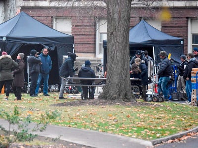 A film crew is gathered on a campus green with people interacting near two black tents. Equipment is visible, and a large tree stands in the foreground. 