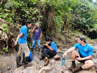Four people in blue shirts are in a lush forest setting. One sits writing on a notepad, another leans in to observe, while the third stands nearby.