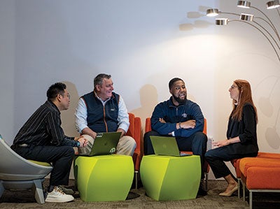 Four people sit in a circle on colorful chairs, engaged in animated conversation, with laptops open. The setting is modern and relaxed.