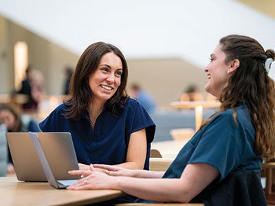 Two women in blue scrubs sit at a table, smiling and talking, with a laptop open in front of them. The setting appears to be a bright, modern space.