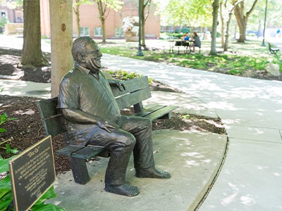 Bronze statue of “Doc Oc” seated on a bench beneath a tree on the Case Western Reserve University campus, overlooking a shaded walkway and green quad. 