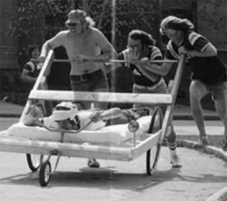 Four students compete in a bed race, with three pushing and one student in a helmet riding