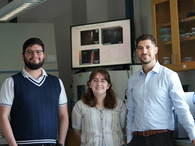 Three researchers stand together in a laboratory, with imaging equipment and a computer display showing material images in the background.