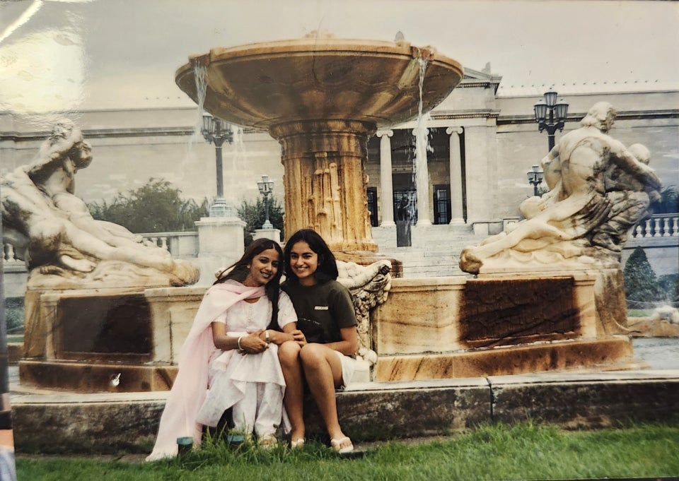Two students, one in a Punjabi dress and one in a t-shirt and shorts, sit on the fountain edge in the Fine Arts Garden with the Museum of Art behind them