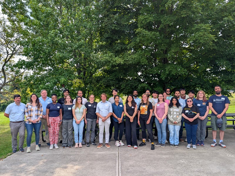 A couple dozen people stand smiling at the camera in an outdoor setting.