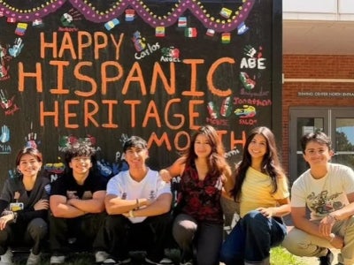 Abigail Figueroa (middle) and friends sit in front of Hispanic Heritage Month sign
