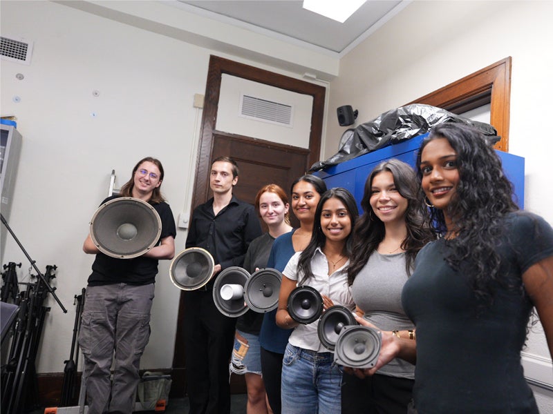 Seven students pose together in a music lab, each holding a speaker of a different size used in the project.