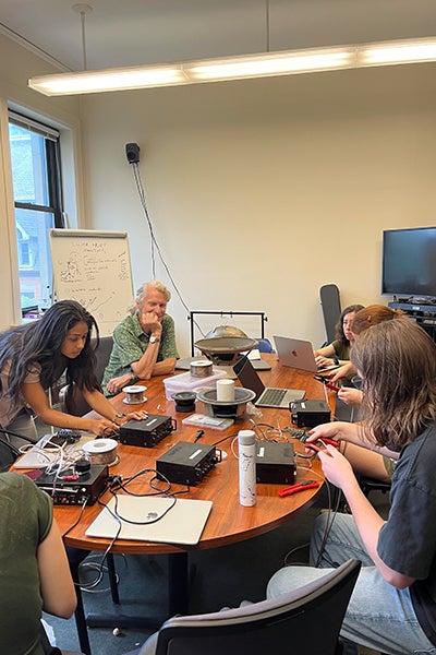 Six people sit around a conference table assembling electronic equipment, with circuit boxes, cables, tools, laptops, and other devices spread across the table.