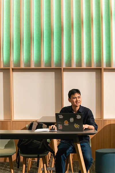 Joshua Kuwana sits alone at a café table with a laptop and notebook, framed by warm wood paneling and glowing green vertical lights.