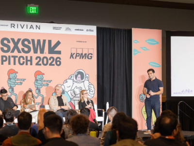 A speaker presents onstage at SXSW Pitch 2026 beside a panel seated beneath a pink event banner, while an audience watches from the foreground.