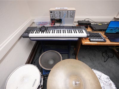 A music lab setup featuring a midi keyboard, electronic audio equipment, a laptop, and mixing controls, with large speakers, a drum, and a cymbal in the foreground.