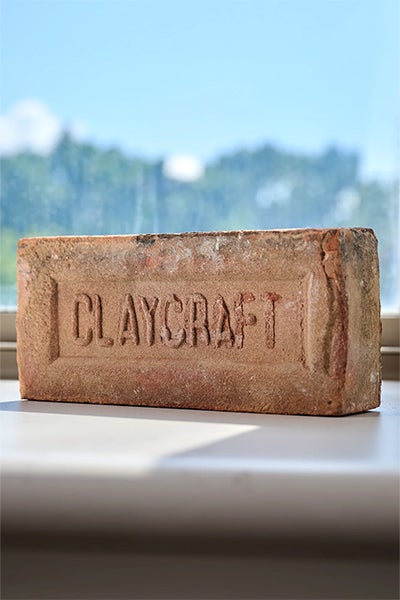 A weathered reddish-brown brick stamped “CLAYCRAFT” rests on a white windowsill, with a softly blurred green landscape and blue sky behind it.