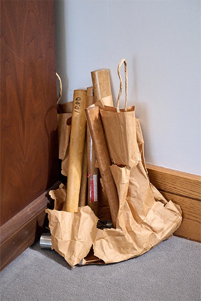 A cluster of wooden handles or tools wrapped in crumpled brown paper leans beside a dark wooden cabinet in a quiet indoor corner.