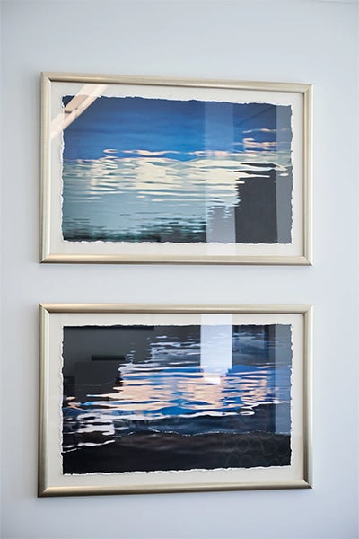 Two framed abstract seascapes hang on a pale wall, their layered blue, black, and white strokes reflecting light and creating a calm, contemplative mood.