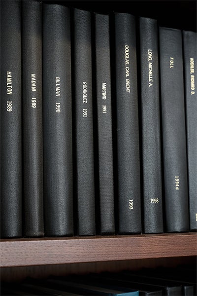 A row of dark clothbound books labeled with authors’ names and dates stands neatly on a wooden shelf, creating a quiet, scholarly library atmosphere.
