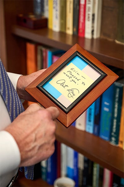 A person holds a wooden frame with a handwritten, signed sticky note in front of a bookshelf. 