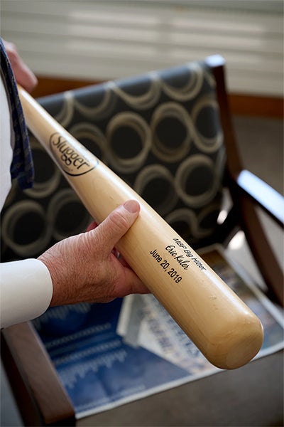 A hand holds a signed Louisville Slugger baseball bat over a patterned chair, with a name and date inscribed on the wood; a quiet, personal keepsake.