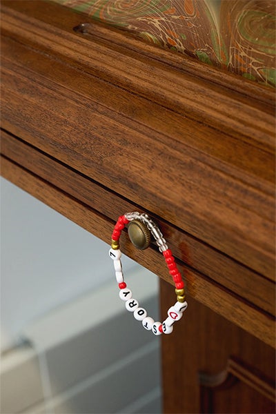 A red-and-white beaded bracelet with letter beads hangs from a brass knob on a wooden drawer, against patterned fabric.