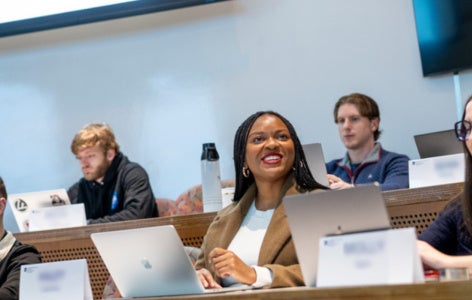 Seven students sit in a tiered classroom in the Peter B. Lewis Building. Two students smile and interact with each other while the others focus on their laptops. 
