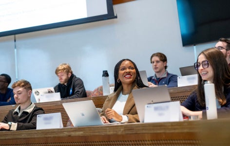 Seven students sit in a tiered classroom in the Peter B. Lewis Building. Two students smile and interact with each other while the others focus on their laptops. 