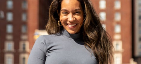 Smiling woman in a gray dress stands outside with tall red brick building in the background. Bright sunlight creates a warm and cheerful atmosphere.