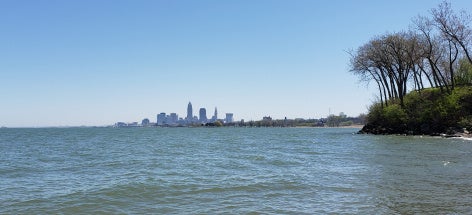 A calm lake with gentle waves is in front of the Cleveland skyline, visible in the distance. A cluster of trees stands on the right shoreline.