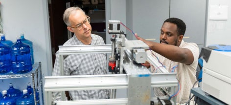 Two men, focused and engaged, work together on a complex machine in a lab. One points attentively while the other observes, discussing adjustments.
