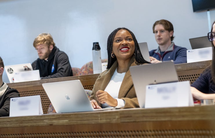 Students in a lecture hall engage with laptops open. A woman in the center smiles attentively, conveying a positive and focused learning atmosphere.