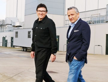 Two men are walking on a concrete surface outside an industrial building. One wears a black jacket, the other a navy blazer and jeans. Both are smiling.