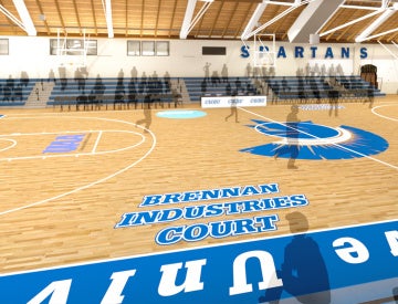 Interior of a gymnasium featuring a basketball court labeled "Brennan Industries Court" with blue and white bleachers and "Spartans" on the wall.