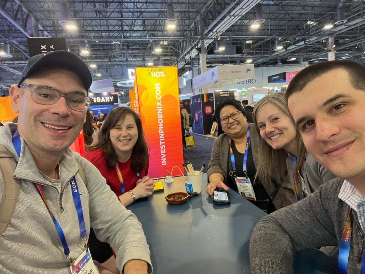 A group of people sit around a table at a busy expo. The setting features bright signage and various booths and banners in the background. 