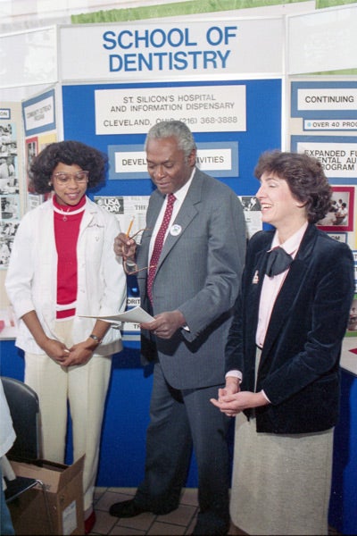 Three people stand smiling in front of a "School of Dentistry" display. The atmosphere is professional and friendly, with educational materials in the background.