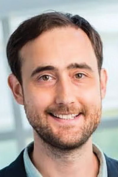 A man with short brown hair and a beard smiles warmly at the camera. He is wearing a light blue shirt and dark jacket, with a blurred background. 
