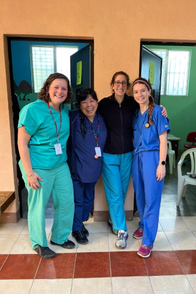 Four people stand in front of a beige wall in scrubs
