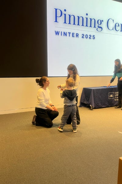 A woman kneels in front of a child before a screen that says Pinning Ceremony.
