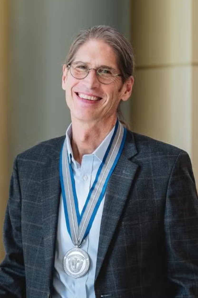 Smiling person with long hair, wearing glasses and a plaid suit, stands with a silver medal around their neck, conveying achievement and pride.