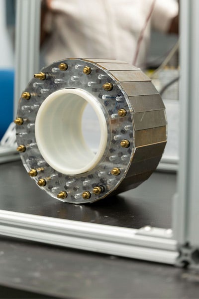 A cylindrical metal object with bolts arranged in a circular pattern on its transparent front, placed on a black surface. Industrial and technical setting.