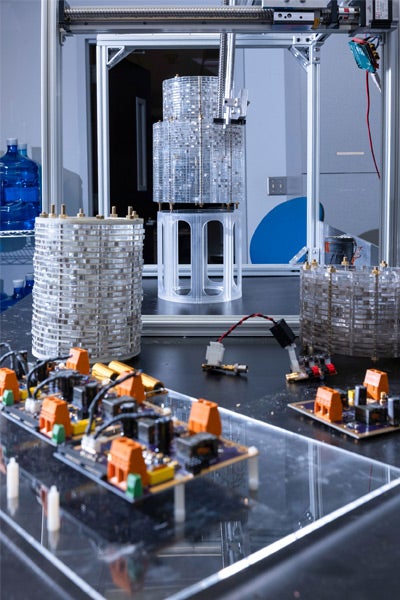 Lab scene with cylindrical transparent structures on a table, surrounded by circuit boards with orange components. The setting appears technical and futuristic.