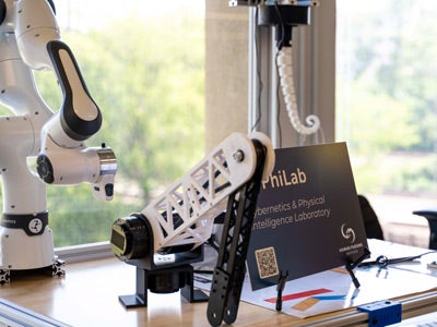 A robot sits on a desk beside a sign, showcasing its friendly design and interactive features in a workspace setting.