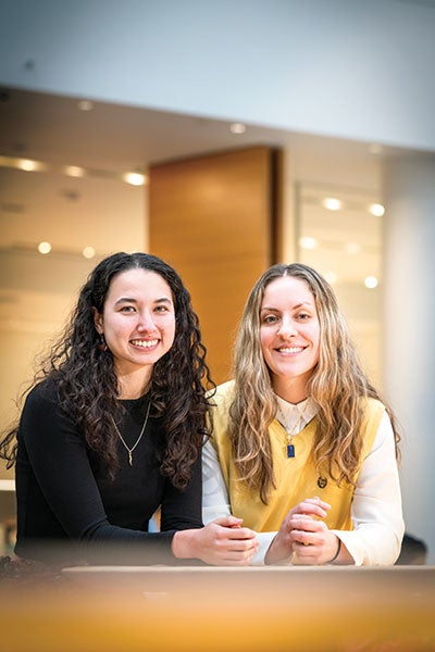 Two women smiling warmly, one in a black top and the other in a yellow vest, pose in a softly lit indoor setting, exuding a friendly and inviting atmosphere.