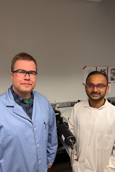 Michael J. A. Hore and Nehal Nupnar stand side by side in a laboratory in front of a light scattering instrument. 