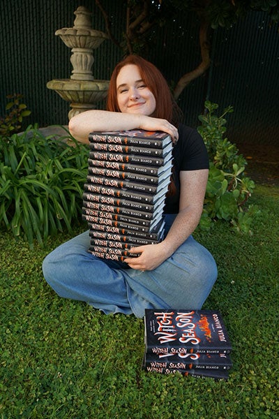 Julia Bianco sitting in the grass with a stack of her new books in her lap