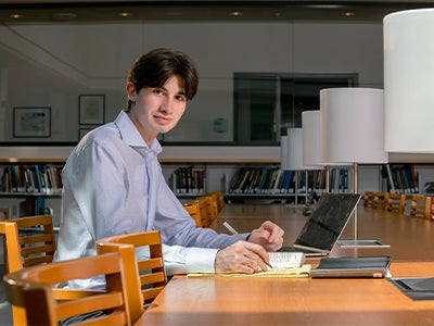 James Calipetro III in a light blue shirt studies at a library table with books and a laptop.