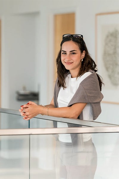 Zeynep Cansev smiles while leaning on a glass railing in a bright, modern interior, conveying a relaxed, welcoming mood.